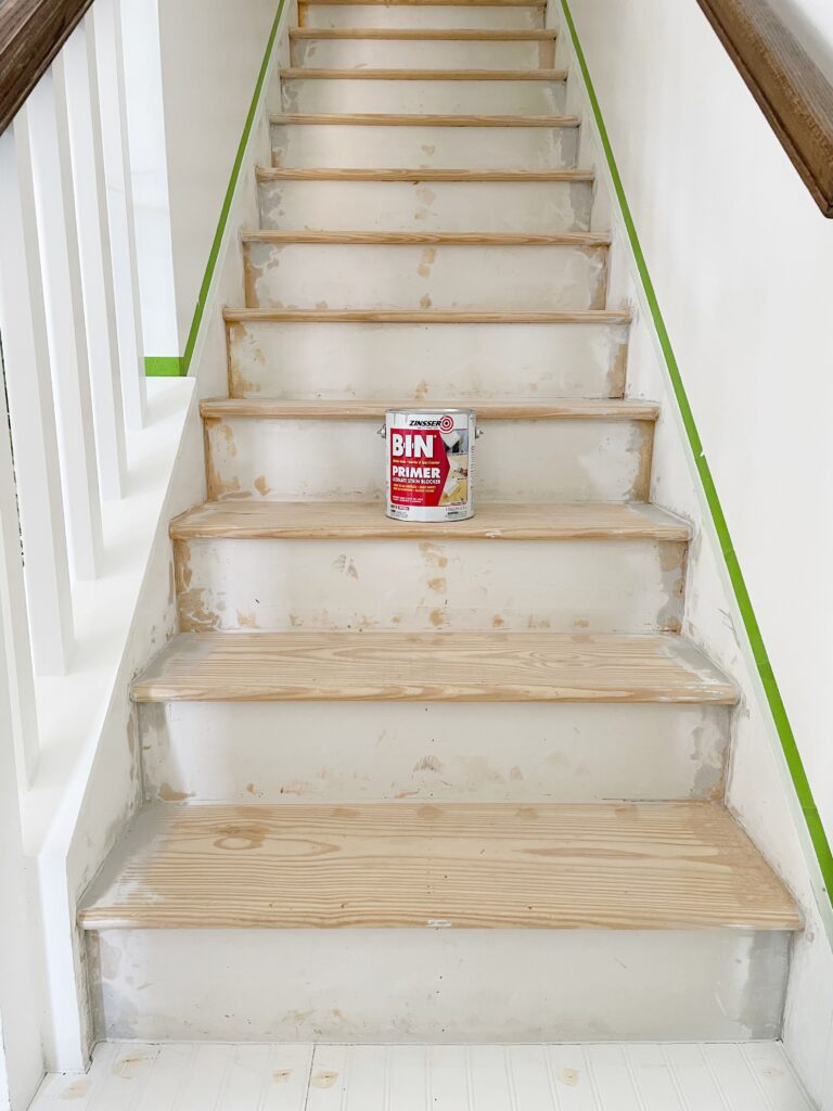 Stripped stairs with a can of BIN primer in the middle. Painter's tape is above the skirt board and goes all the way up the stairs