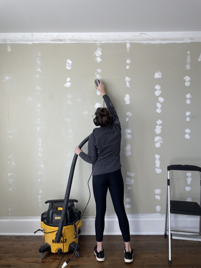 Elisha from Our Aesthetic Abode is sanding down a wall that's been patched with joint compound. She's using a mouse sander connected to a shop vac with a PVC adapter.
