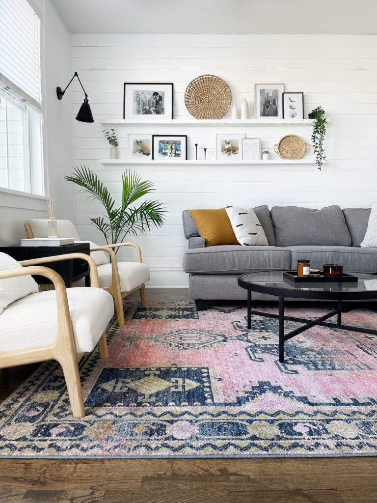 A white living room with a horizontal shiplap wall, a gray couch, a pink bohemian style rug, black coffee table, and a lot of decor. 