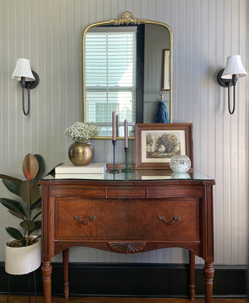 A view of Elisha from Our Aesthetic Abode's beadboard entryway makeover featuring a vintage vanity, some plants, a brass vase, black sconces, vintage art, black candlesticks, and a crystal jar