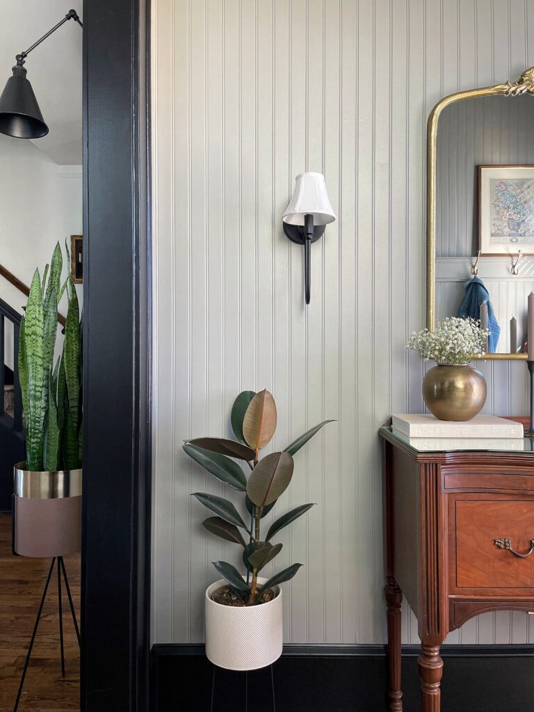 A view of Elisha from Our Aesthetic Abode's beadboard entryway makeover featuring a vintage vanity, some plants, a brass vase, and a black sconce.