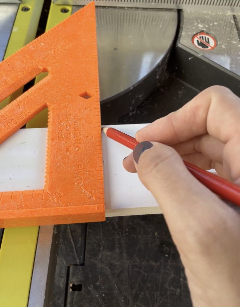 Elisha from Our Aesthetic Abode is demonstrating how to use a speed square which is #7 on her beginner DIYer checklist. She has it attached to a board and is drawing a line against the straight edge.