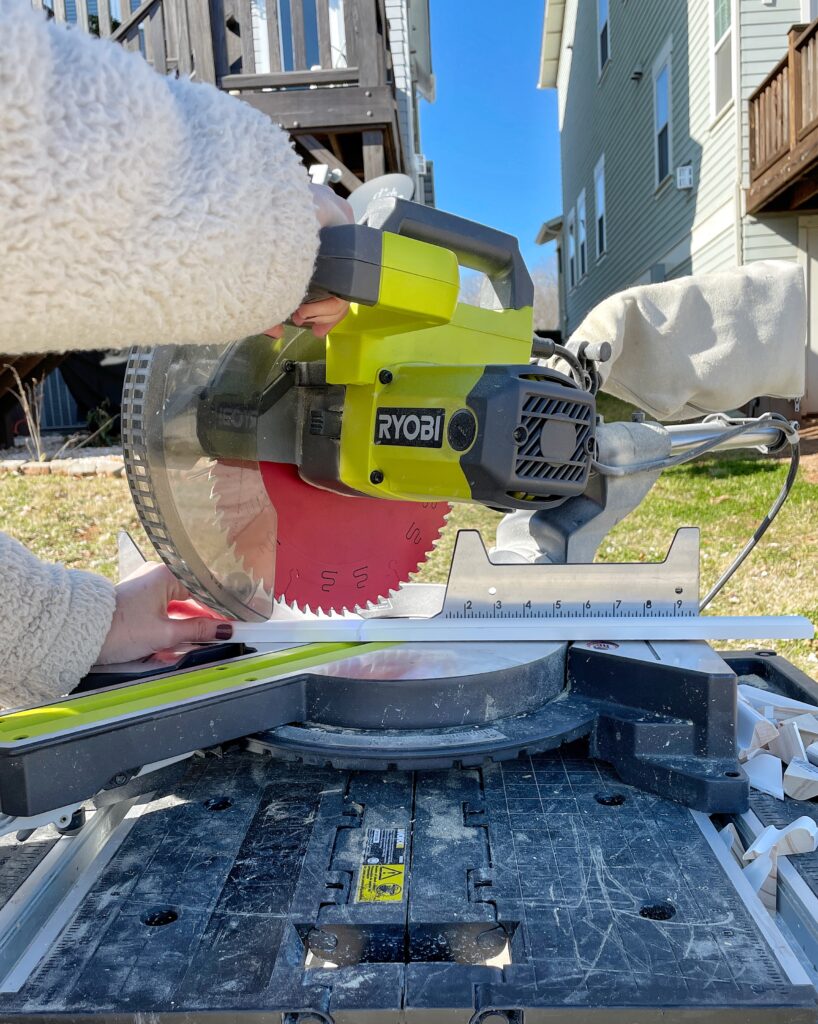 Elisha from Our Aesthetic Abode is using her miter saw to cut an angle on her trim piece for her Recessed Wainscoting Accent Wall. The miter saw is casting a shadow on the wood where the blade will cut through.