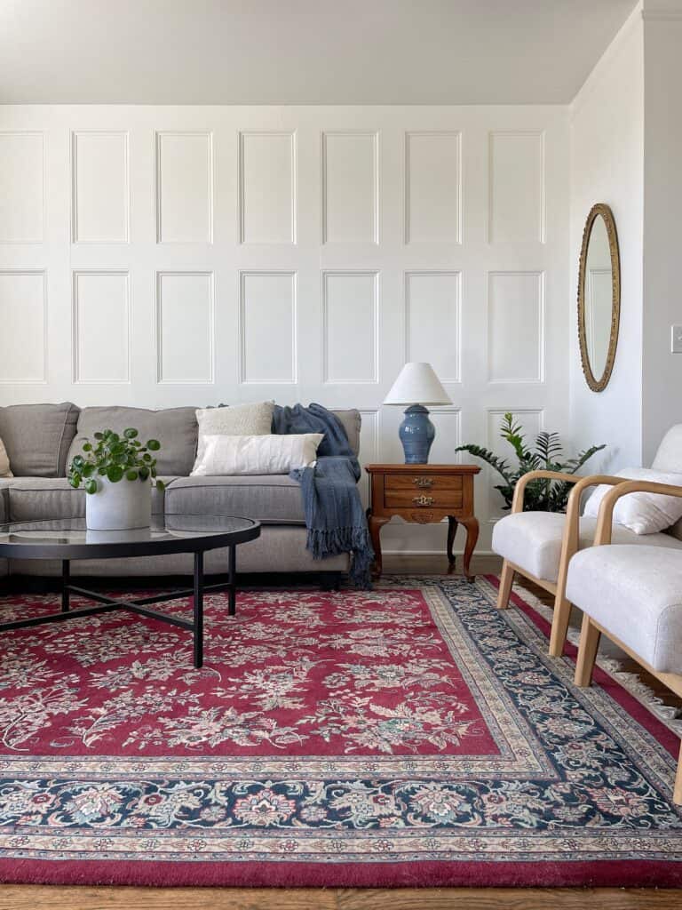 Elisha from Our Aesthetic Abode's completed DIY recessed wainscoting accent wall in her furnished living room with a gray couch, a beautiful red oriental rug, and two beige upholstered accent chairs.